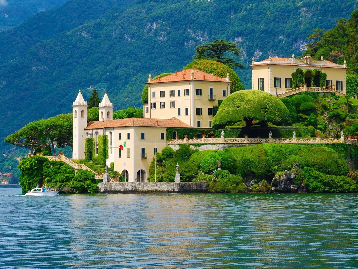 Prachtvolle historische Villa mit weissen Türmen, üppigen Terrassengärten und gestutzten Bäumen direkt am Seeufer, umgeben von bewaldeten Bergen im Hintergrund und einem Boot auf dem ruhigen Wasser