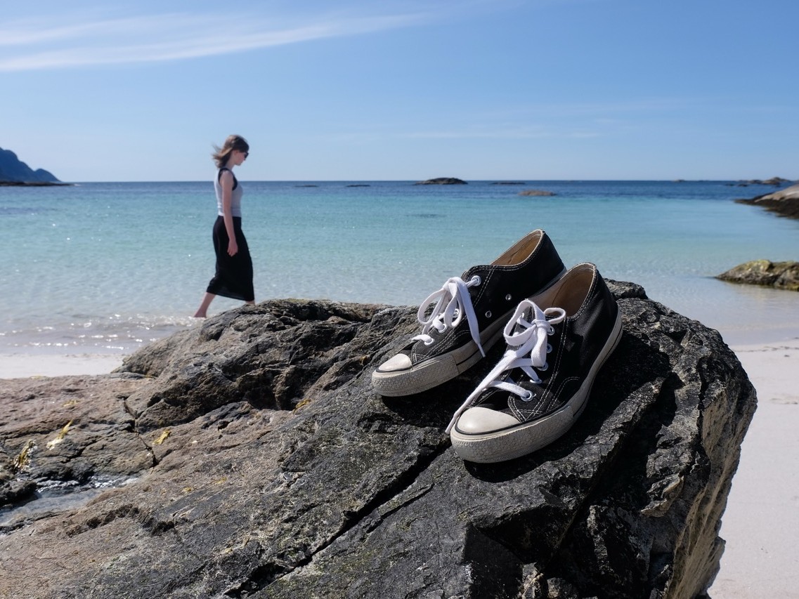 Schwarze Sneakers auf einem Felsen im Vordergrund, im Hintergrund eine Frau die barfuss an einem türkisblauen Sandstrand entlangläuft