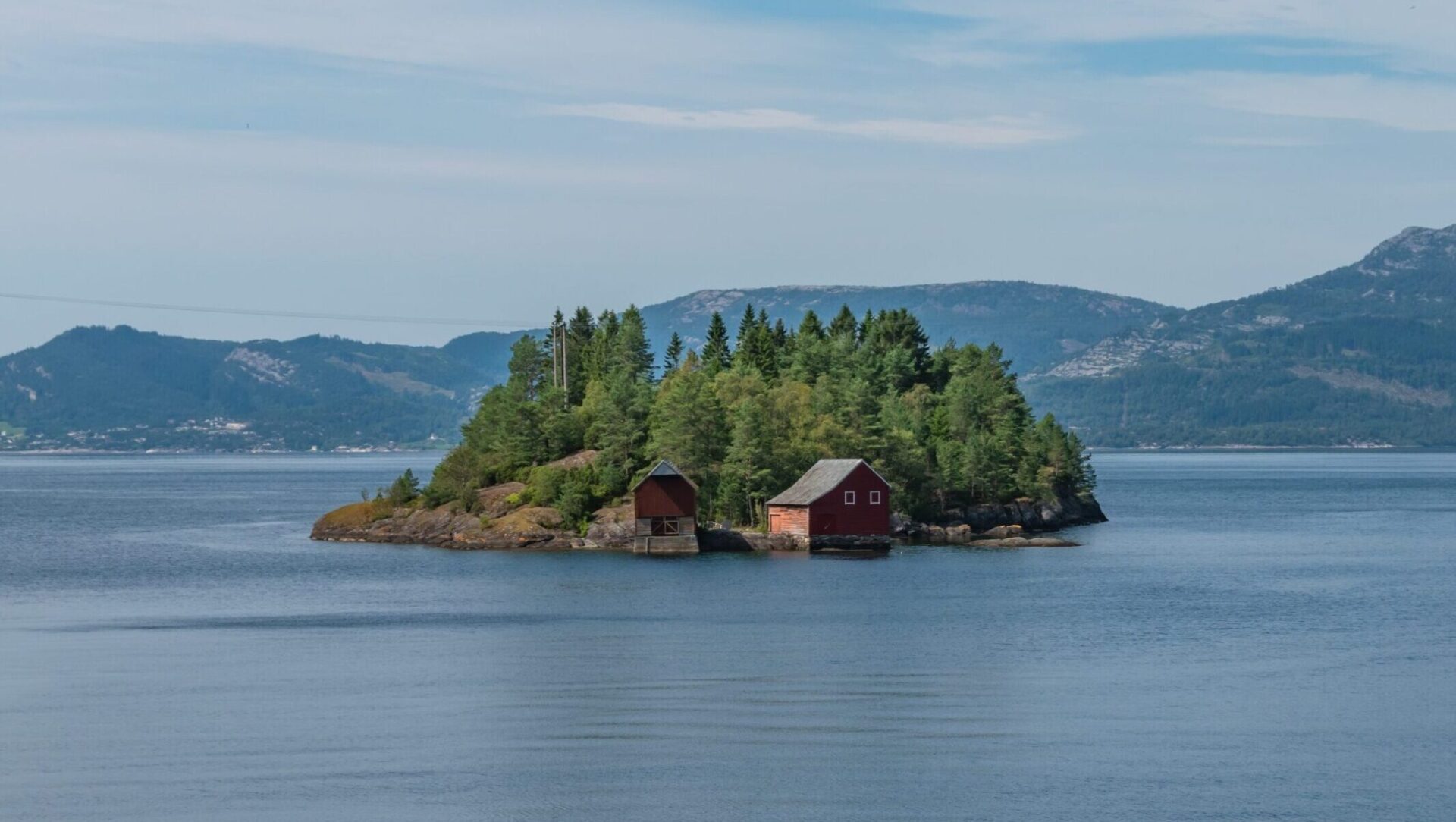 Kleine bewaldete Insel mit zwei roten Holzhütten am Ufer, eingebettet in einen ruhigen norwegischen Fjord mit Bergen im Hintergrund – Privatinsel Lysfjord, 2.2 Hektar, zum Verkauf über ISLA AURUM